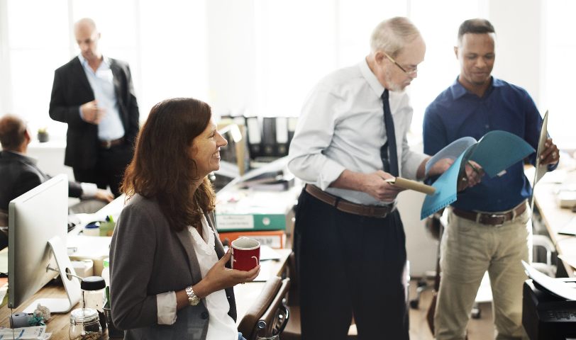 Two men stand together looking at a report while a woman holding coffee sits beside them
