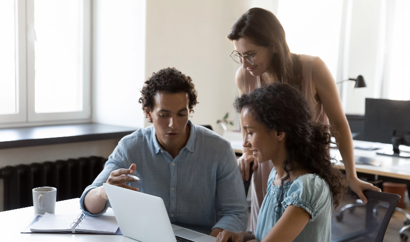 A group of three people stand around a computer and discuss